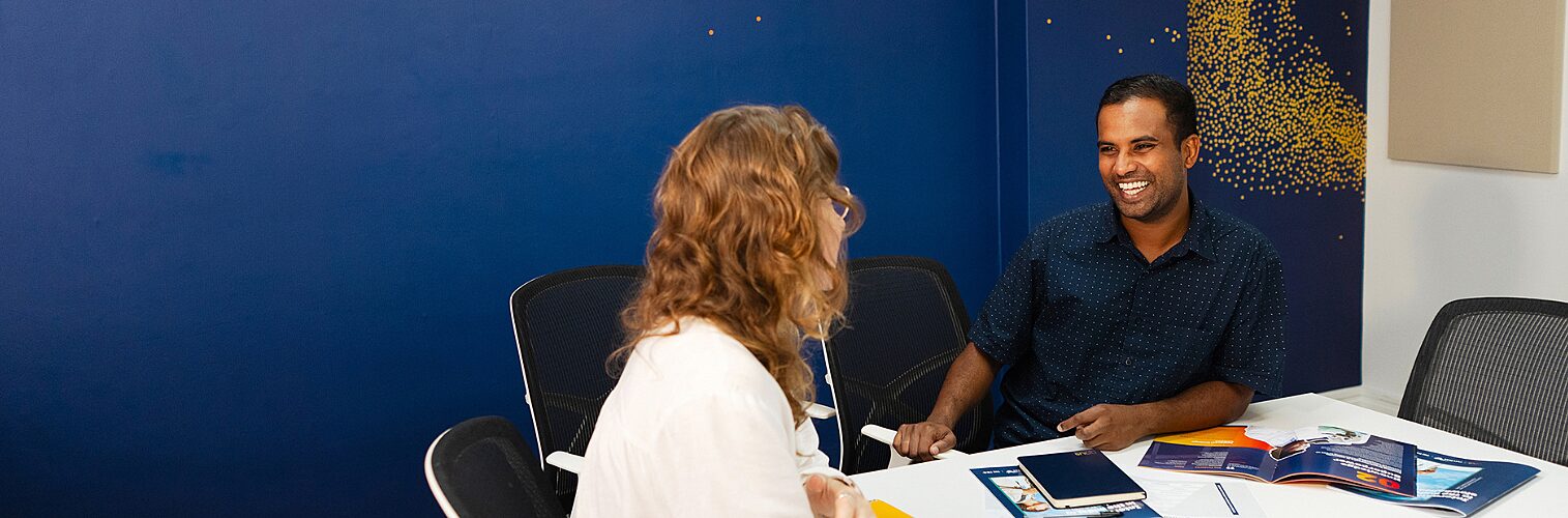 Nous UK Director, Minto Felix sits at a table in a meeting room with a colleague, engaged in discussion. MInto Felix faces the camera while his colleague is seen from behind. Papers, a notebook, and an open booklet are spread across the table, and a dark blue wall with a yellow patterned design forms the background.