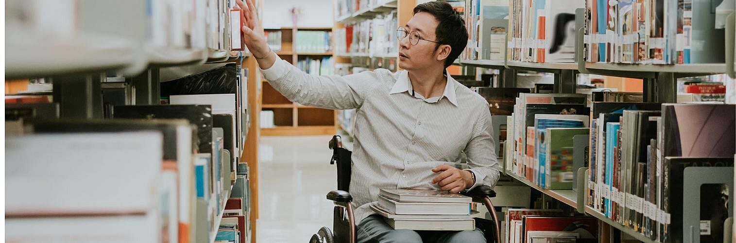 Disabled student in a wheelchair perusing books at a library.