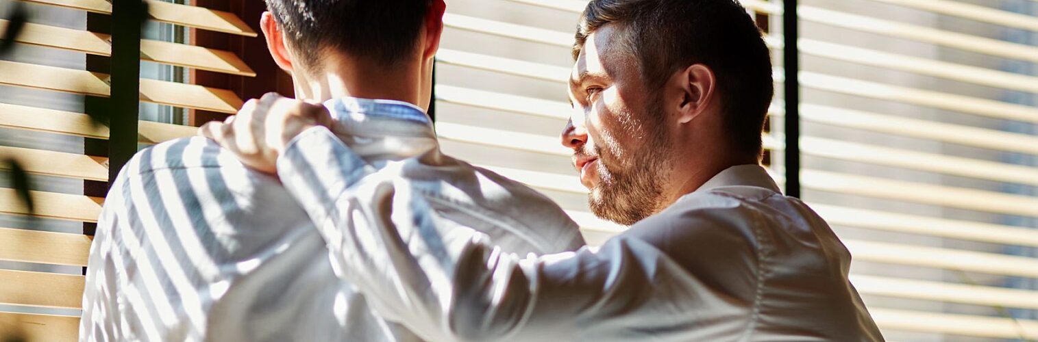 Two young businessmen supporting one another near a window,