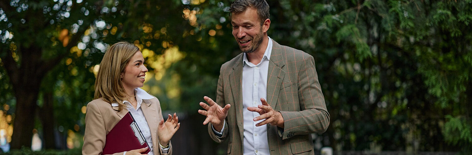 Two colleagues walking and talking outdoors, discussing ideas during an informal meeting in a green urban setting.