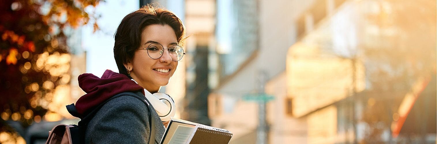 A young female university student standing with books and smiling at the camera.