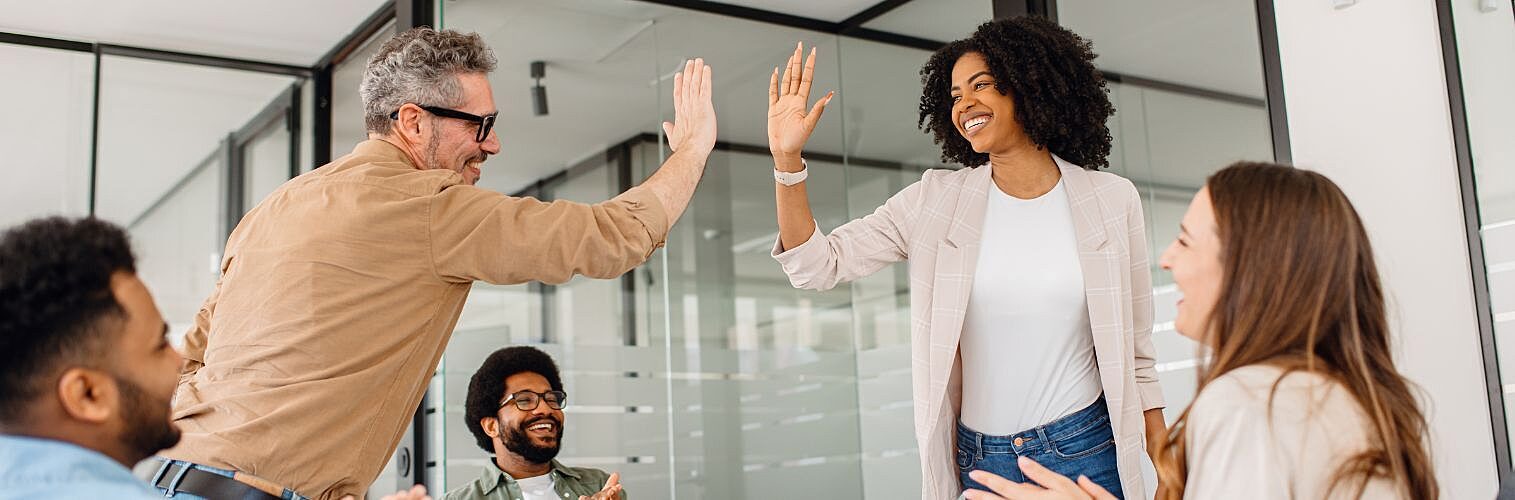 A diverse team of professionals celebrates a workplace achievement with a high-five in a modern office.