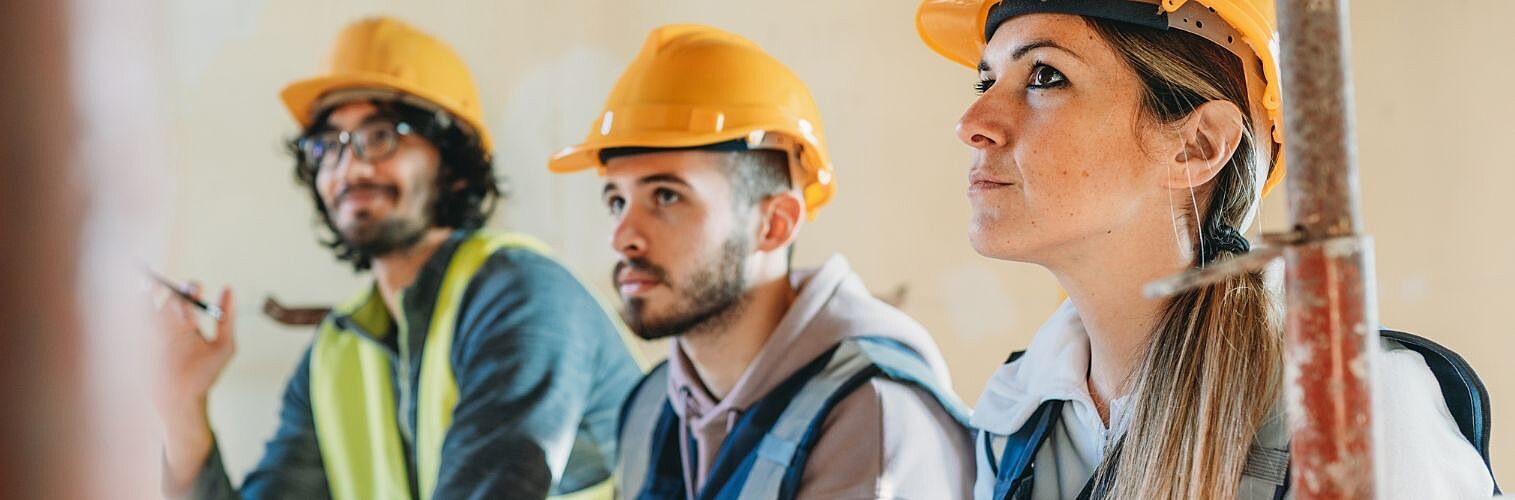 Three people are listening to their foreman on a construction site. They are sitting on the worksite.