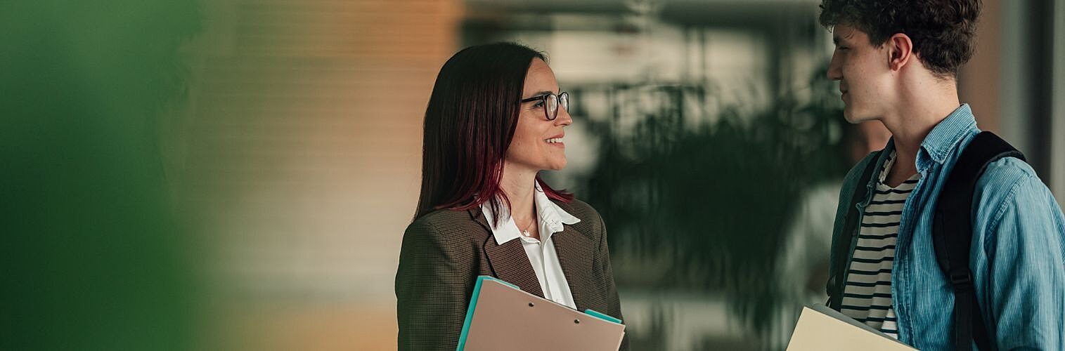 Female teacher and young male student having a candid discussion and sharing insights while standing in a modern school hallway.