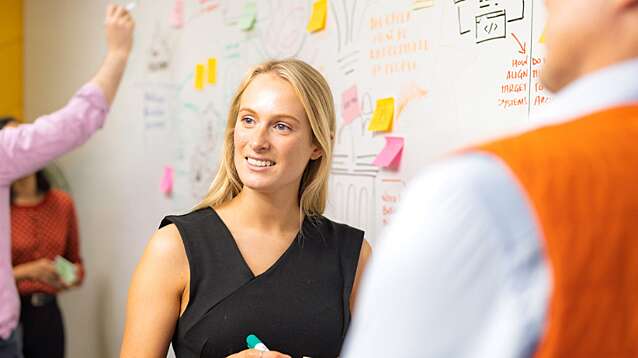 A small group of people collaborate in an office meeting area, standing in front of a large whiteboard covered with notes, sketches, and colourful sticky notes. One person in the centre holds a marker and faces others while two more people are partially visible in the foreground. Another person in the background writes on the board. The space is brightly lit, with a yellow wall visible on the left and a busy, creative planning setup on the whiteboard.